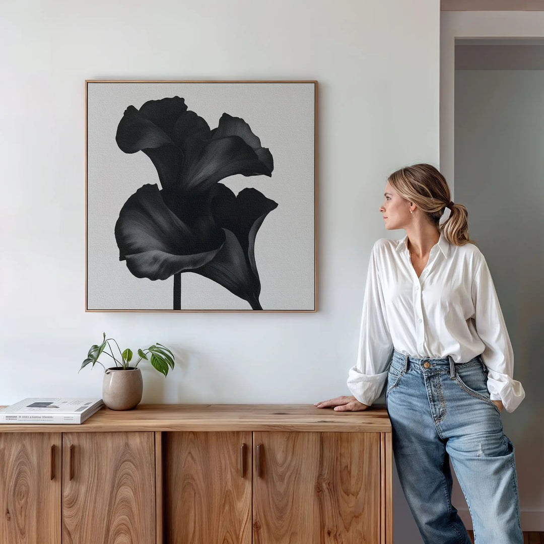 A woman in a white blouse and jeans leans against a wooden sideboard, admiring the Noir Bloom Canvas Art - Square, a striking minimalist piece, displayed in a modern living room with a potted plant and books.