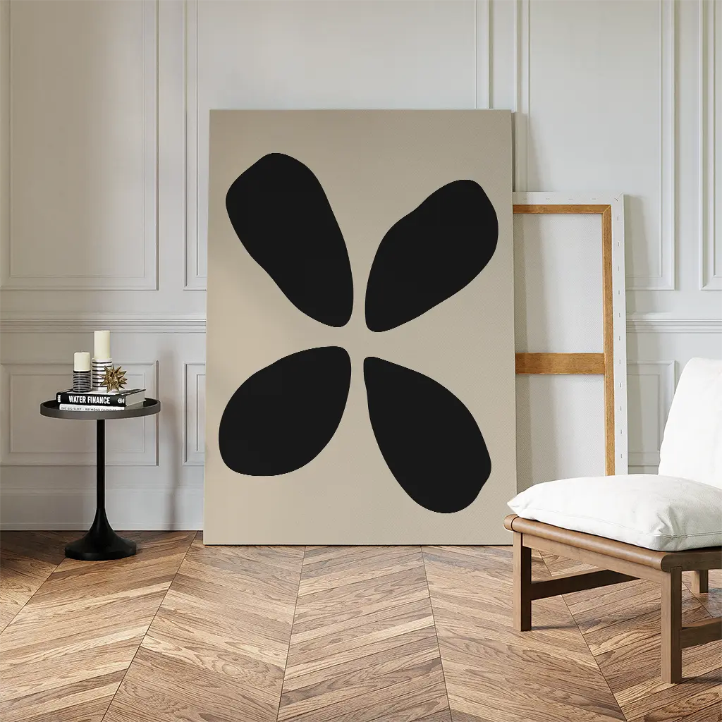 A minimalist room with modern decor features Noir Botanica Canvas Art—four black petal shapes on beige—leaning against the wall beside an empty frame, a chair, and a small round table with candles and books.