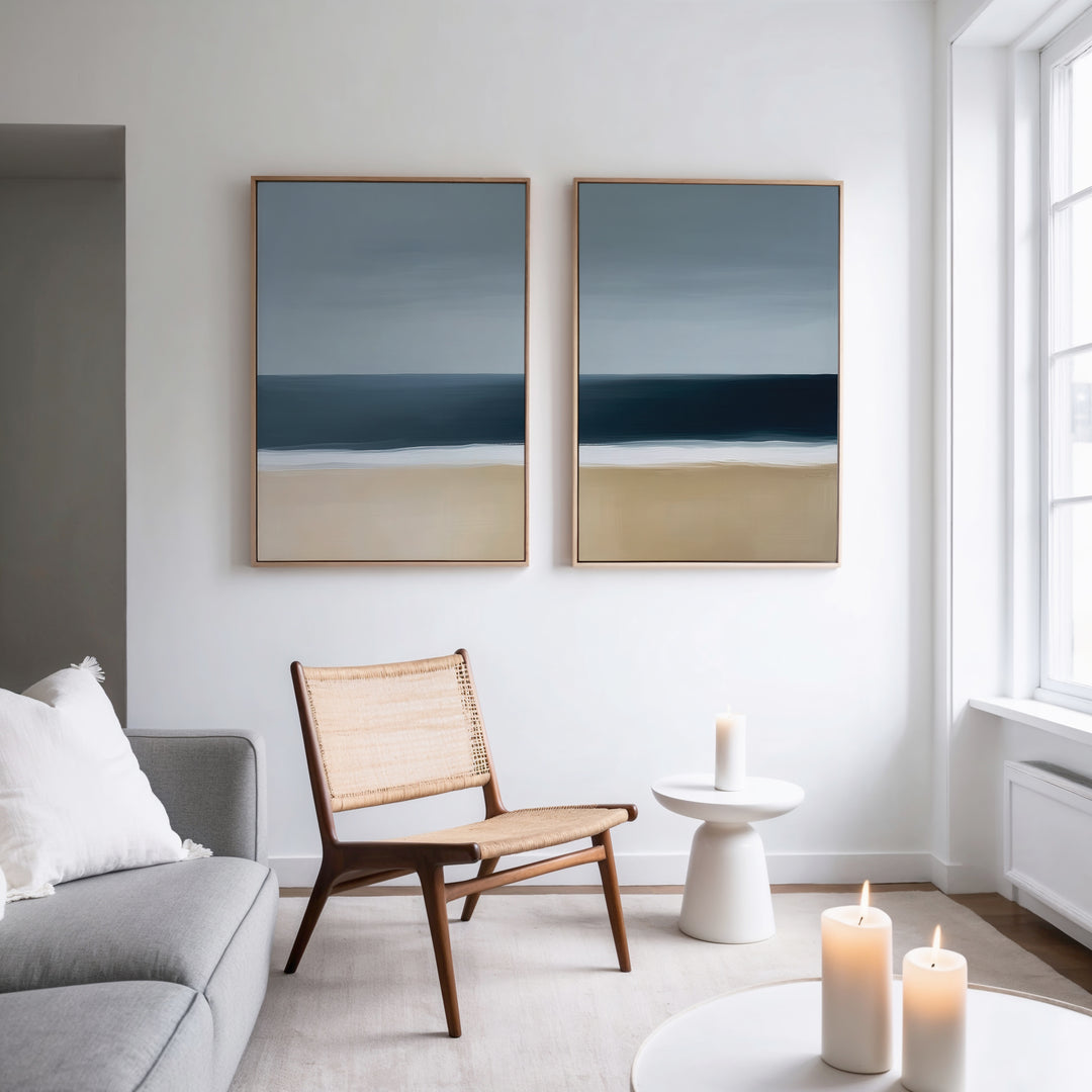 A modern, minimalist living room features the Ocean Edge Set of 2 woven chairs, a light gray sofa, and a small round white table with a candle. Abstract art above and three candles by the large window complete this perfect living room decor.