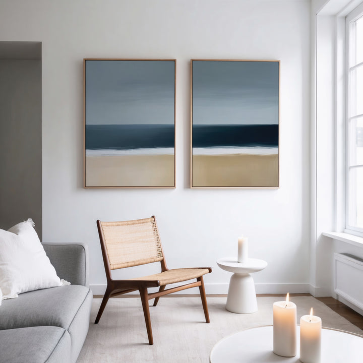 A modern, minimalist living room features the Ocean Edge Set of 2 woven chairs, a light gray sofa, and a small round white table with a candle. Abstract art above and three candles by the large window complete this perfect living room decor.