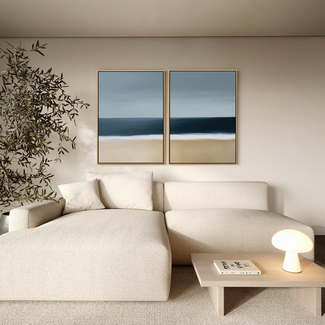 A modern living room with a cream sectional sofa, wooden coffee table, indoor plant, and the Ocean Edge Set of 2 coastal wall art above the couch adds a touch of elegant decor.