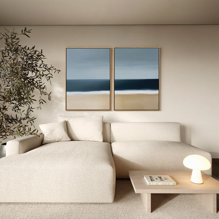 A modern living room with a cream sectional sofa, wooden coffee table, indoor plant, and the Ocean Edge Set of 2 coastal wall art above the couch adds a touch of elegant decor.