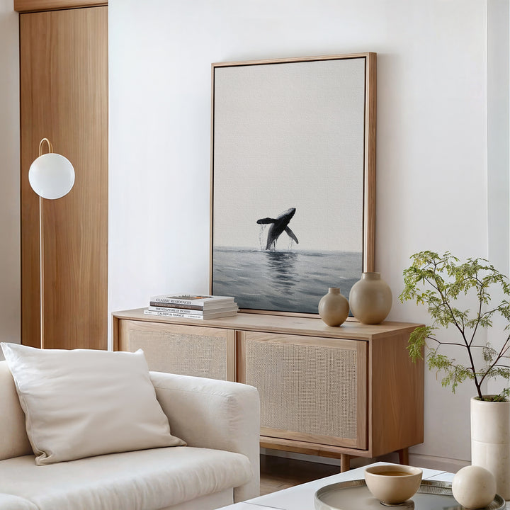 A minimalist living room with neutral tones, a white sofa, light wood cabinet, ceramic vases, a small plant, and the Ocean Leap Canvas Art featuring a jumping whale over the ocean.