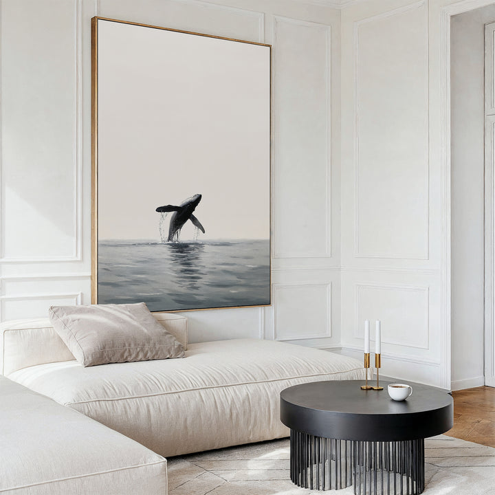 A modern living room with a cream sofa, round black coffee table, two candlesticks, and the striking Ocean Leap Canvas Art—a large framed painting of a breaching whale—hanging on a white paneled wall.