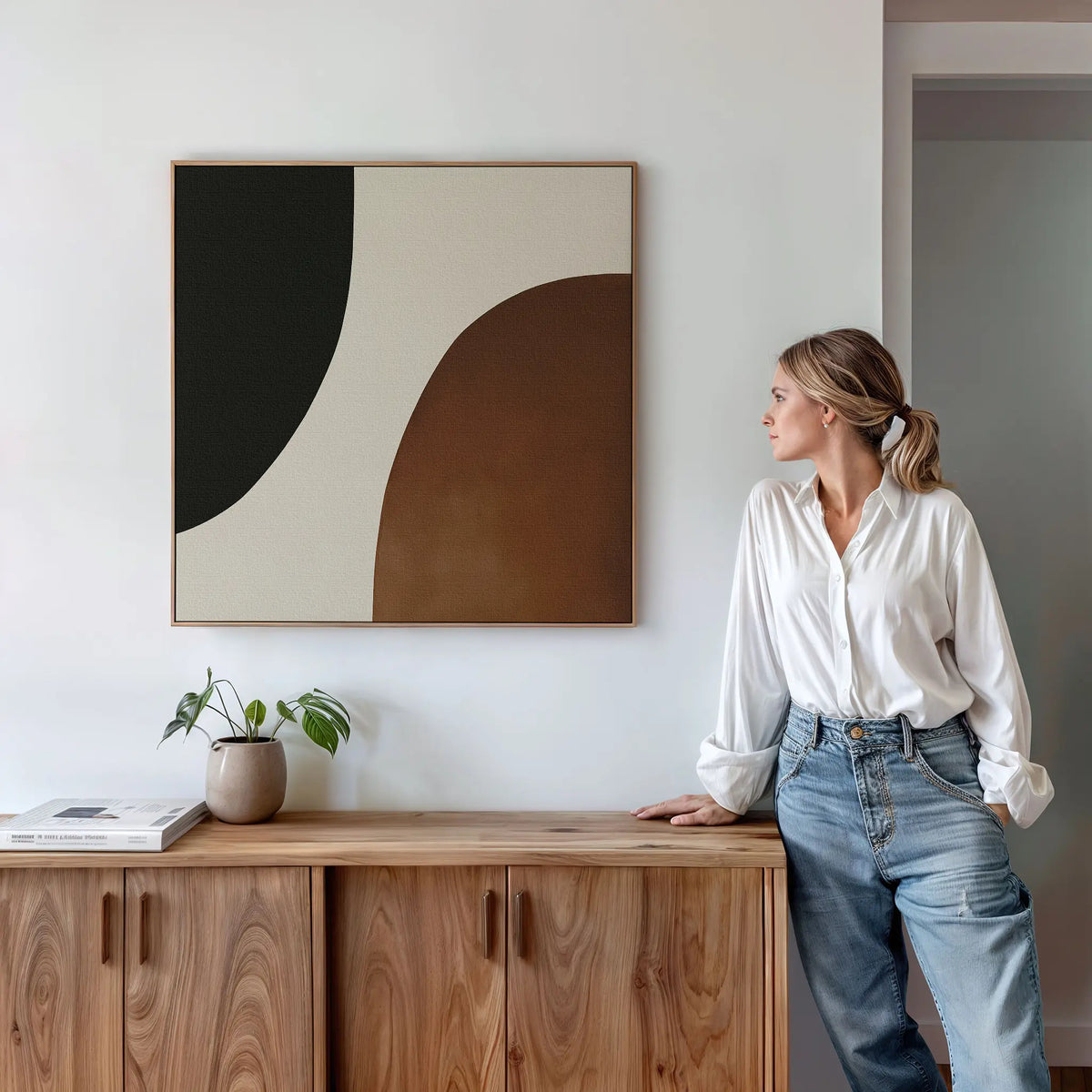 A woman in a white shirt and blue jeans leans against a wooden cabinet, admiring the Organic Balance Canvas Art—modern abstract wall art with black, white, and brown shapes—in a bright, minimalist living room.