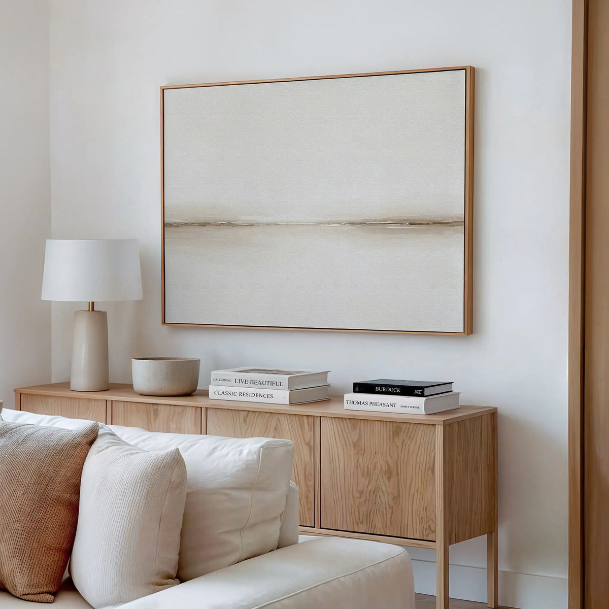 A minimalist living room with a wooden sideboard, neutral decor, books, a white table lamp, a bowl, and the Pale Desert Canvas Art in beige tones above a light-colored sofa—perfect for stylish living room decor.
