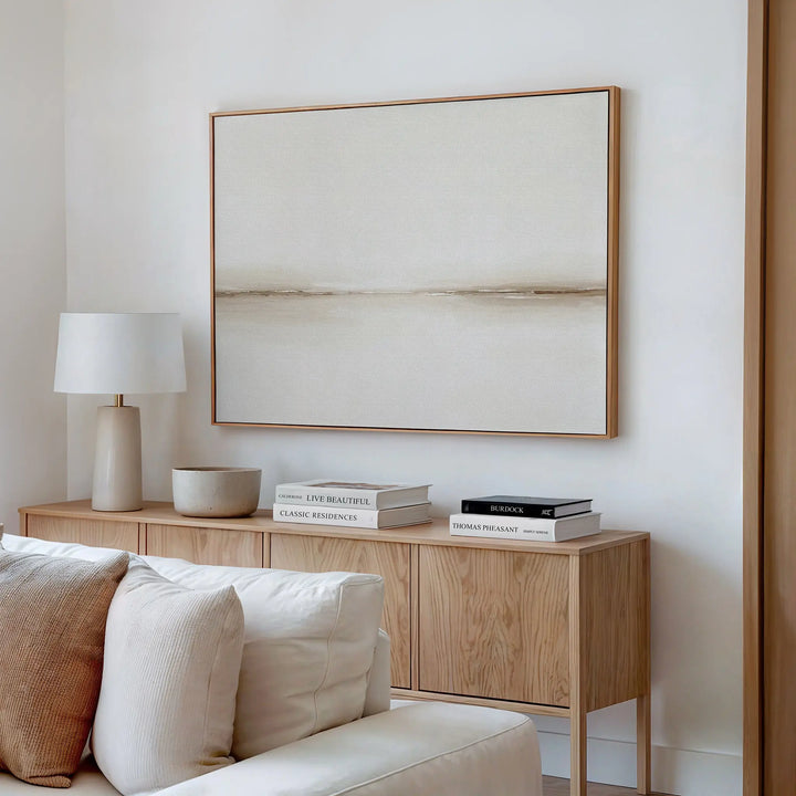 A minimalist living room with a wooden sideboard, neutral decor, books, a white table lamp, a bowl, and the Pale Desert Canvas Art in beige tones above a light-colored sofa—perfect for stylish living room decor.