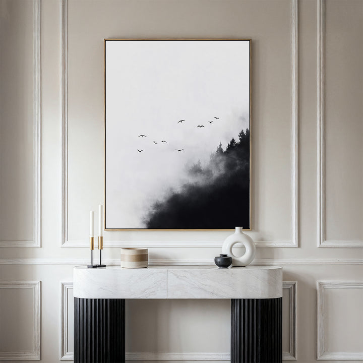 A modern console table with minimalist decor stands against a beige paneled wall, topped by the Passing Pines Canvas Art—a large piece showing bird silhouettes flying above misty black trees.