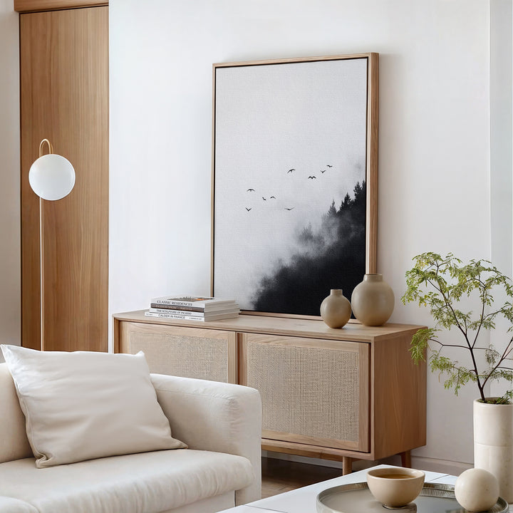 A minimalist living room with light wood furniture, a white couch with beige cushions, ceramic vases, a small green plant, and the Passing Pines Canvas Art—a striking wall art piece featuring trees and birds on a foggy backdrop.