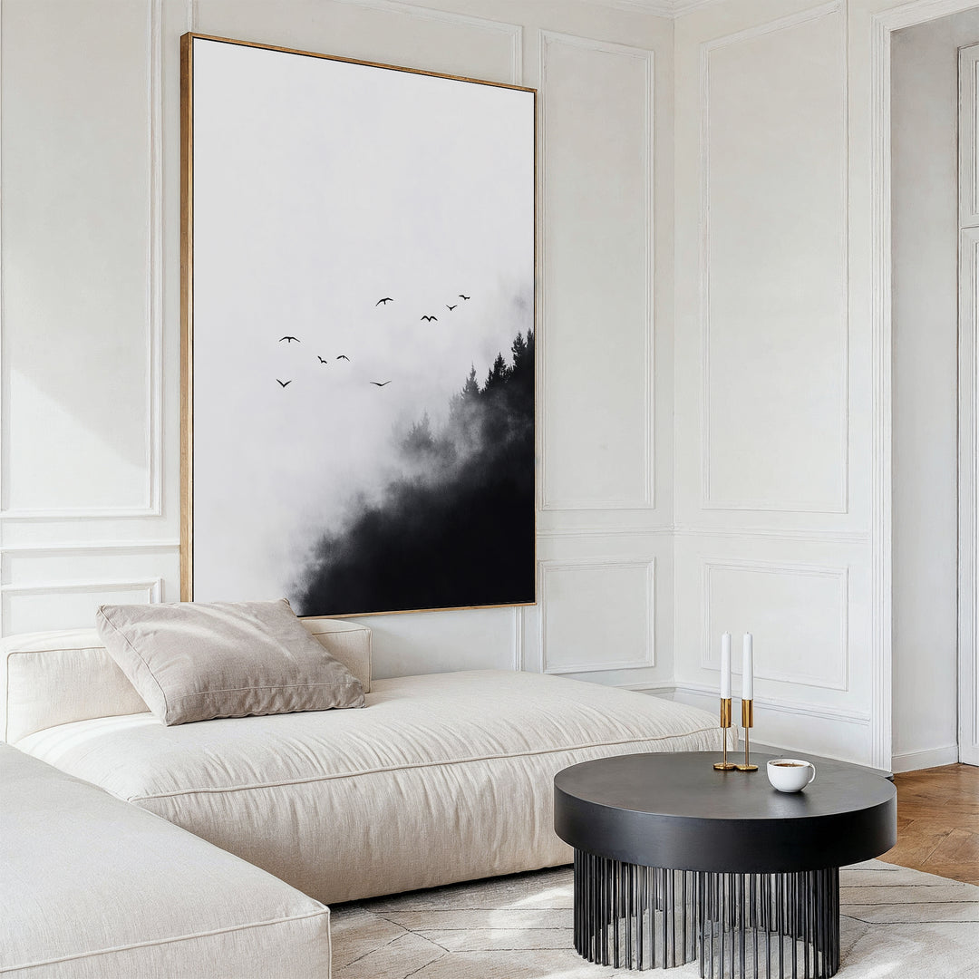 A minimalist living room featuring a white sectional sofa, beige cushion, round black coffee table with candles and a cup, complemented by the Passing Pines Canvas Art—showing birds soaring above misty trees.
