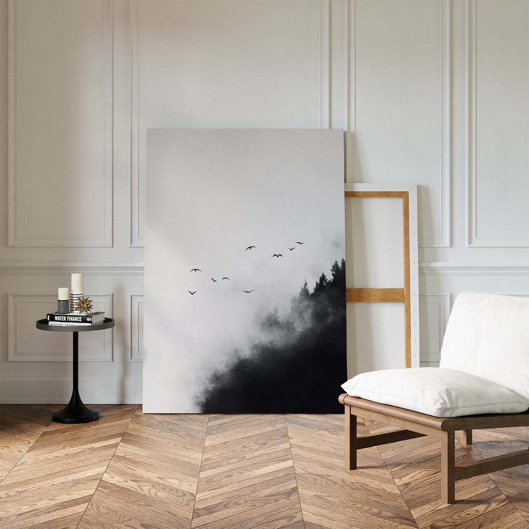 A minimal room with wood herringbone floors, white paneled walls, a cushioned chair, black side table with candles, and the Passing Pines Canvas Art—large wall decor of misty trees and birds—resting against the wall.