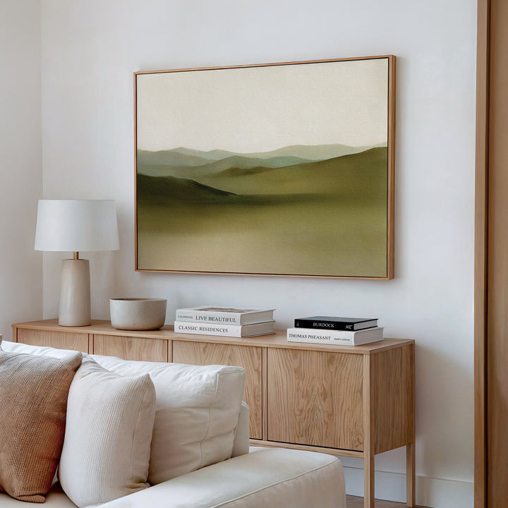 A modern living room with a light wood sideboard, books, a white lamp, and beige couch features minimalist decor and serene wall art—the Pastoral Peace Canvas Art, a large abstract landscape painting with green hills—for an inviting touch.