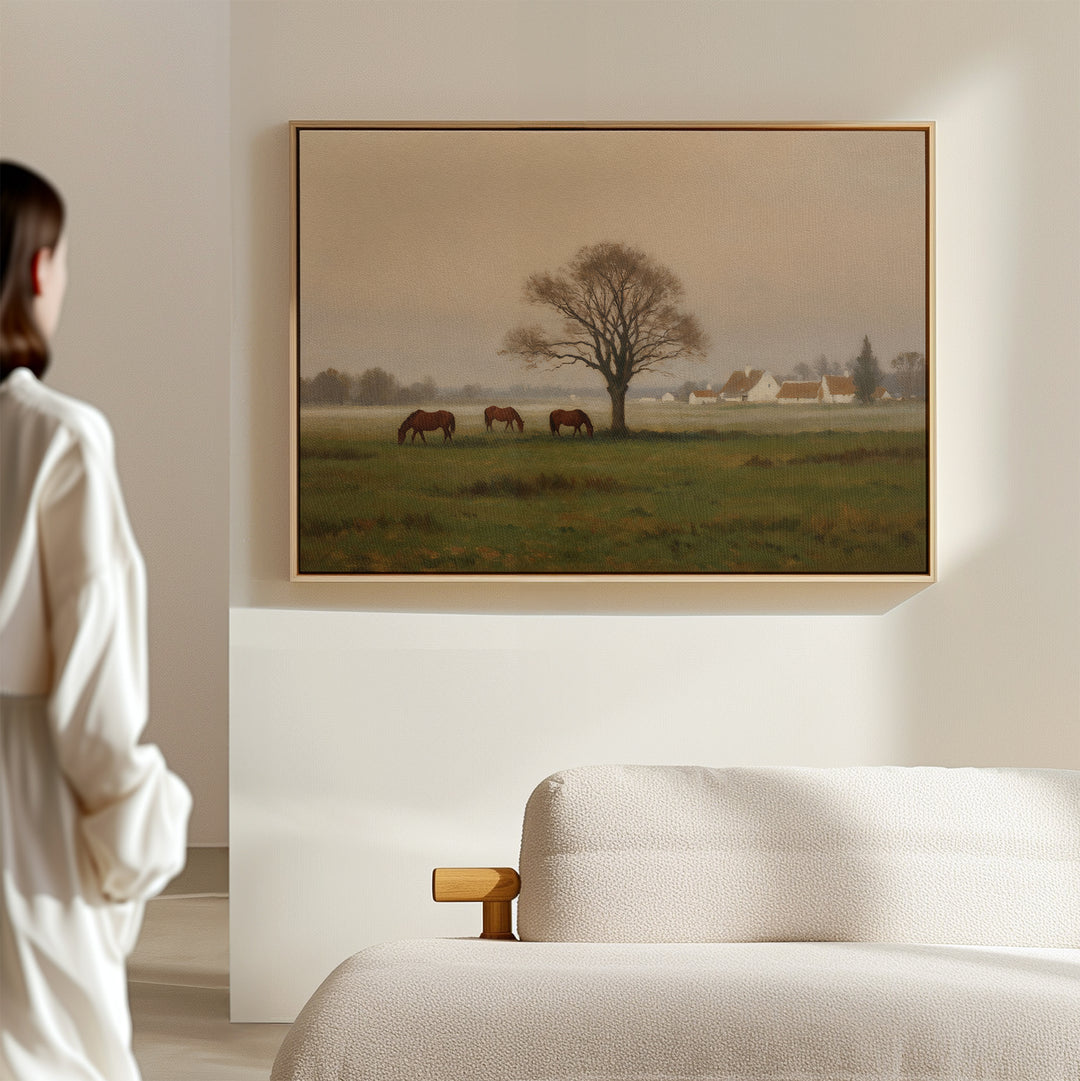 In a minimal, softly lit room, someone admires the Pasture Morning Canvas Art, which shows horses grazing near a tree in a tranquil rural scene with quaint houses in the distant countryside.