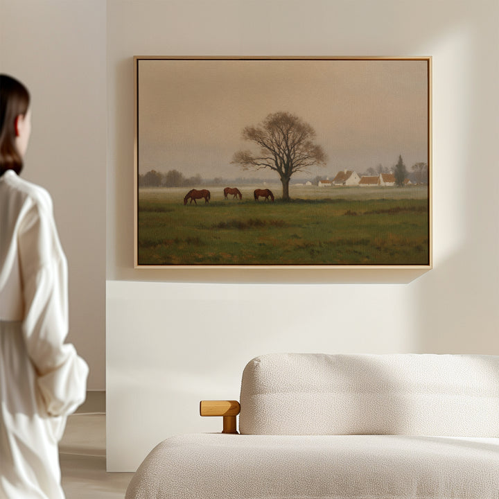 In a minimal, softly lit room, someone admires the Pasture Morning Canvas Art, which shows horses grazing near a tree in a tranquil rural scene with quaint houses in the distant countryside.