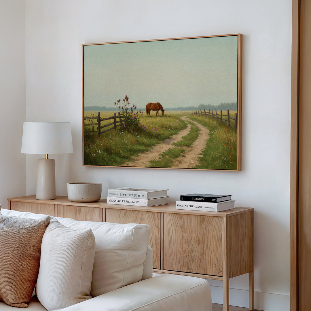 A cozy living room features a light wood sideboard with stacked books, a white lamp, and a bowl. Above hangs the Pasture Path Canvas Art, depicting a horse grazing near a fence along a curving country path in a grassy field.