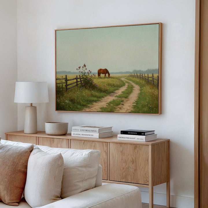 A cozy living room features a light wood sideboard with stacked books, a white lamp, and a bowl. Above hangs the Pasture Path Canvas Art, depicting a horse grazing near a fence along a curving country path in a grassy field.