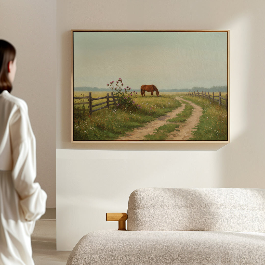 A person in a white shirt stands in a softly lit modern room, admiring the Pasture Path Canvas Art—a framed painting on the wall showing a horse grazing by a dirt path and wooden fences in the countryside.