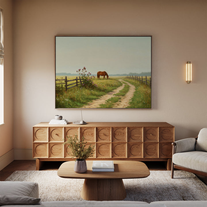 A cozy living room with neutral tones features a wooden credenza, a coffee table with a vase, a beige sofa, and rustic wall art—a large Pasture Path Canvas Art depicting a horse grazing near a fence on a country path.