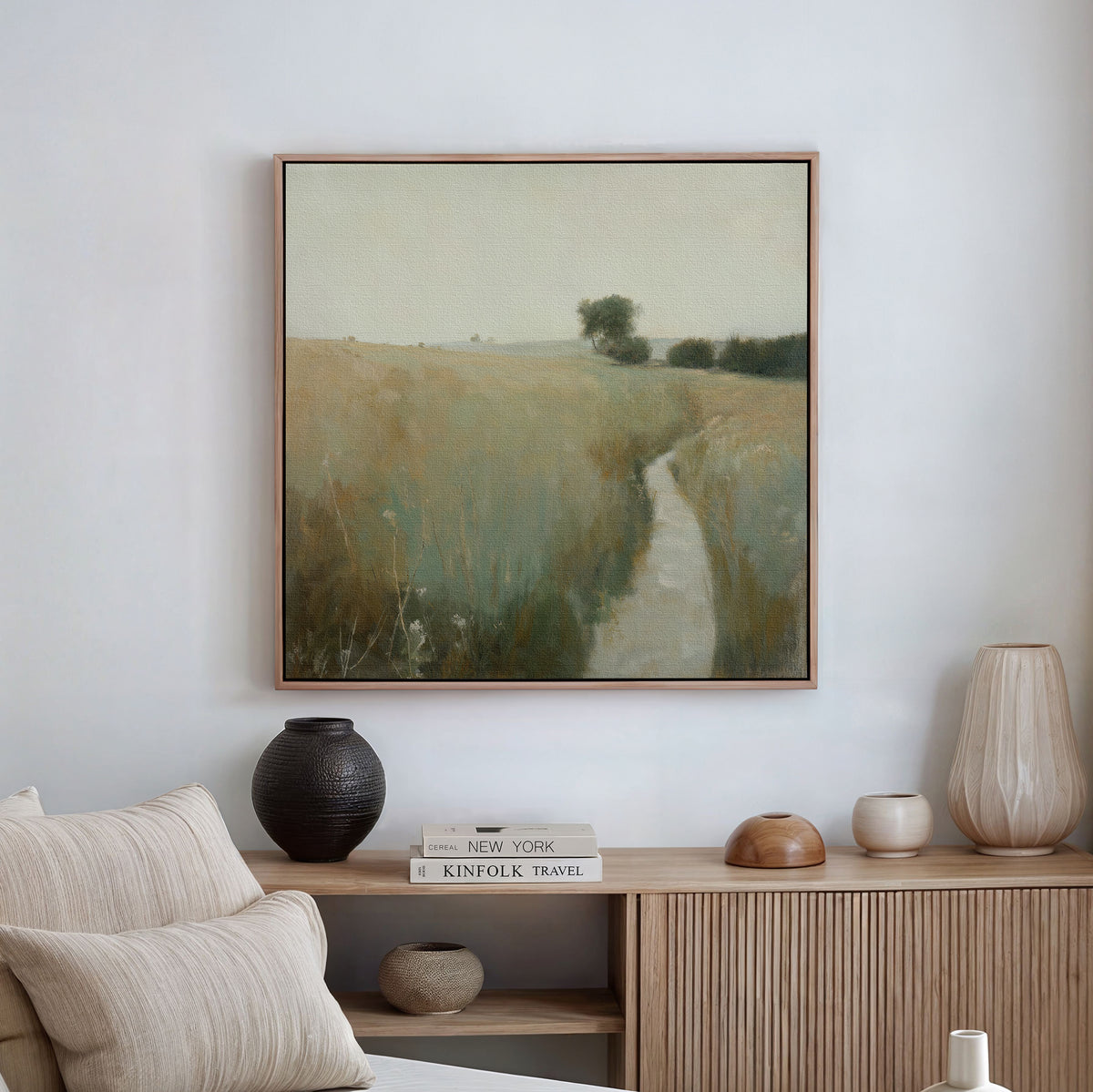 A serene living room with beige tones features rustic decor and the Path Through the Meadow Canvas Art. Ceramic vases and books rest on a wooden cabinet, while a light sofa invites nature lovers to relax.