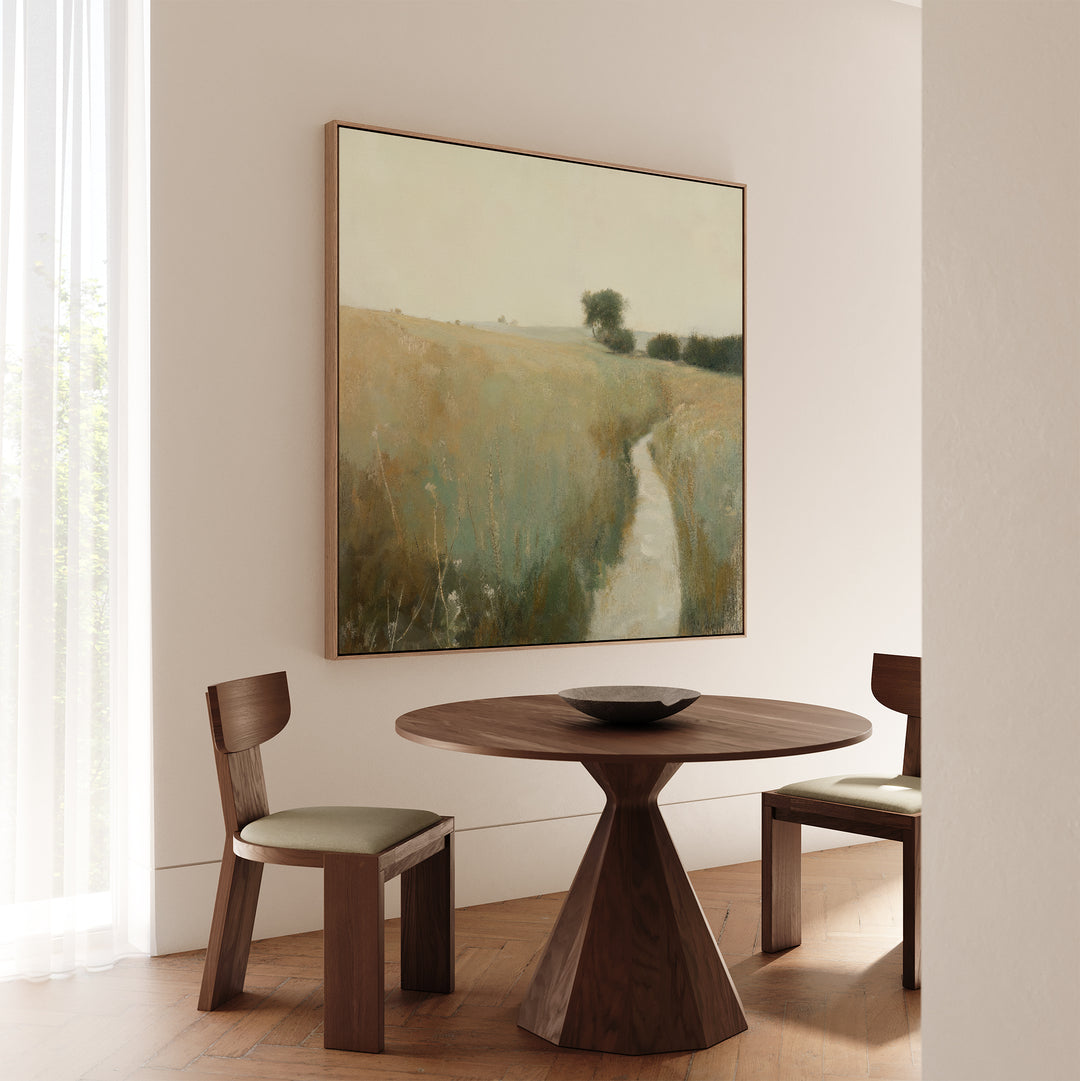 A minimalist dining area features a round wooden table, two cushioned chairs, and rustic decor with the Path Through the Meadow Canvas Art landscape. Soft natural light through sheer curtains creates a serene setting for nature lovers.