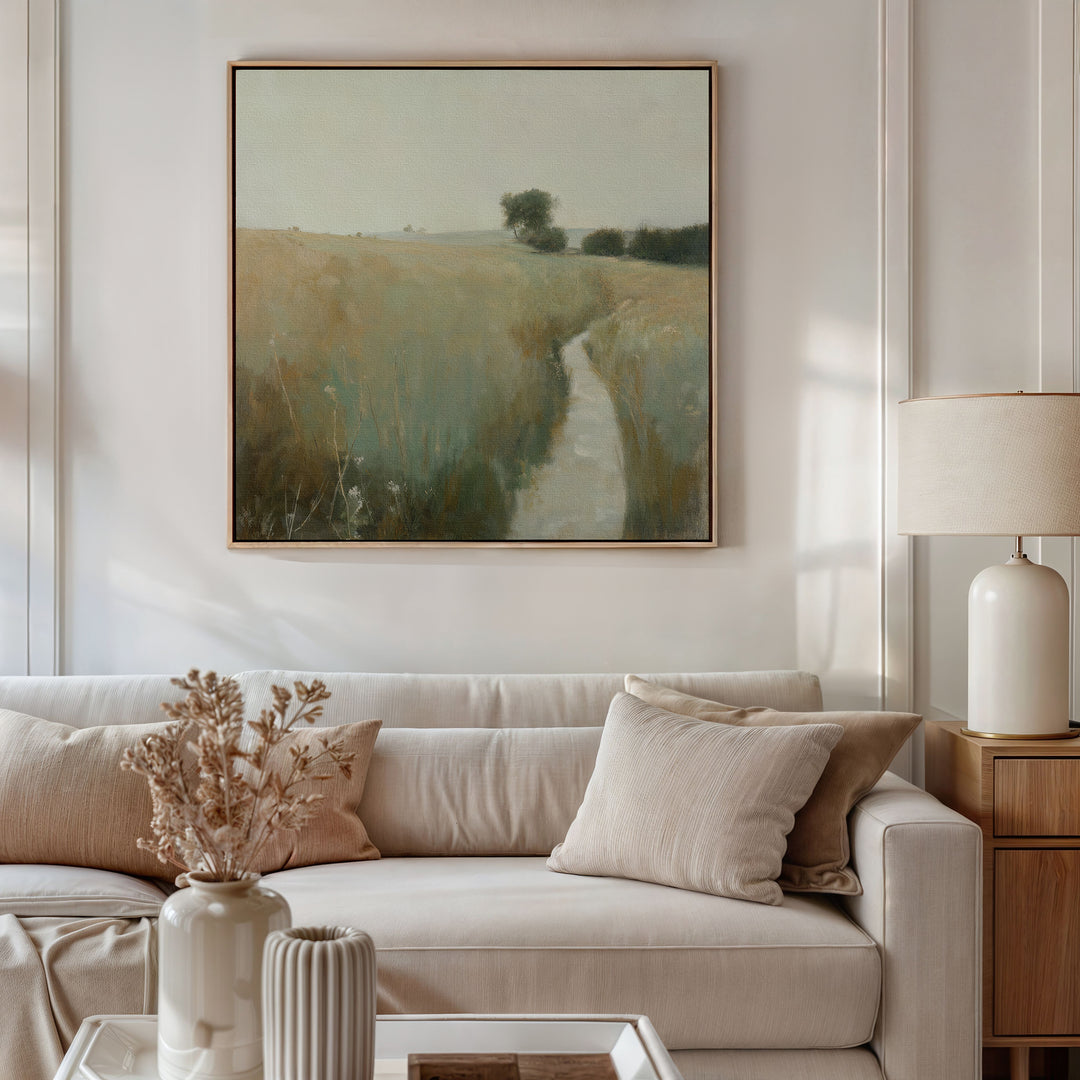 A cozy living room with a beige sofa, neutral pillows, and rustic decor features a side table with a lamp, dried flowers, and Path Through the Meadow Canvas Art—a landscape painting perfect for nature lovers.