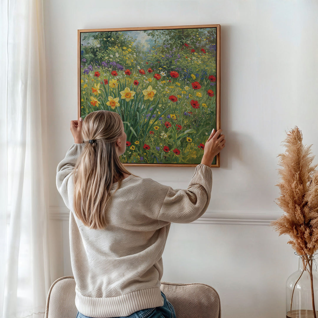 A woman with blonde hair in a ponytail hangs the Poppy Meadow Canvas Art, a vintage-inspired wildflower print, on a white wall. Dried pampas grass accents the corner, and soft natural light filters through a sheer curtain.