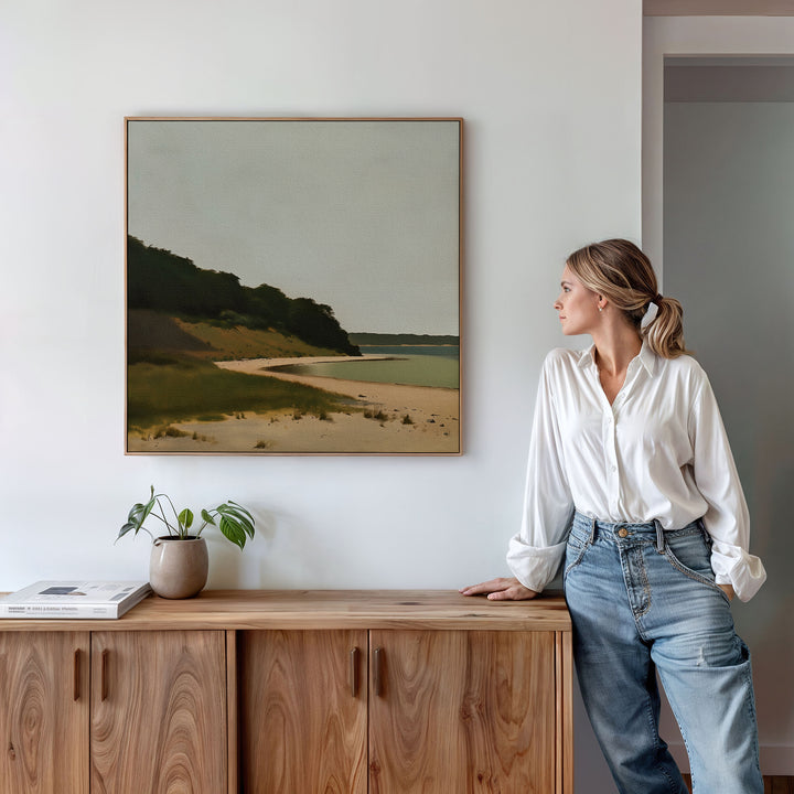 A woman in a white blouse and jeans leans against a wooden cabinet, admiring the Quiet Bend Canvas Art—a minimalist landscape painting that adds elegance to her living room. A small plant and books rest on the cabinet.
