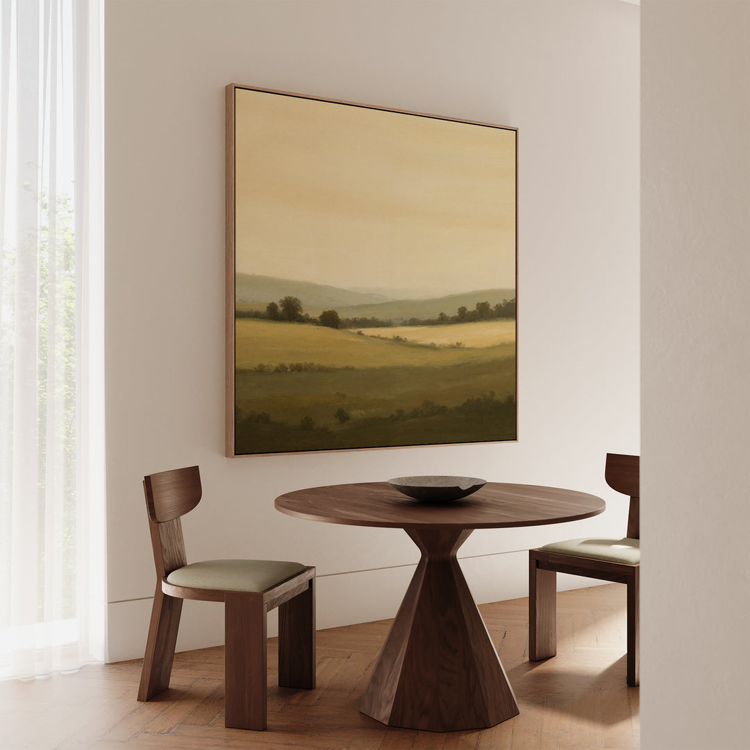 A minimal dining space with a round wooden table, two cushioned chairs, a shallow bowl centerpiece, and the Quiet Countryside Canvas Art—a large landscape painting—brings rural landscape decor to life.