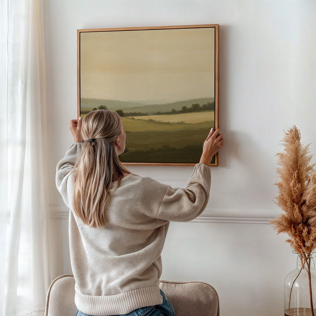 A woman with blonde hair in a ponytail hangs the Quiet Countryside Canvas Art on a white wall in a softly lit room decorated with sheer curtains and a vase of dried pampas grass.