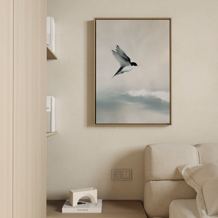 The Quiet Flight Canvas Art, featuring a bird soaring above clouds, is displayed on a beige wall above a light sofa in a minimalist living room—ideal for those who love serene, modern wall decor.