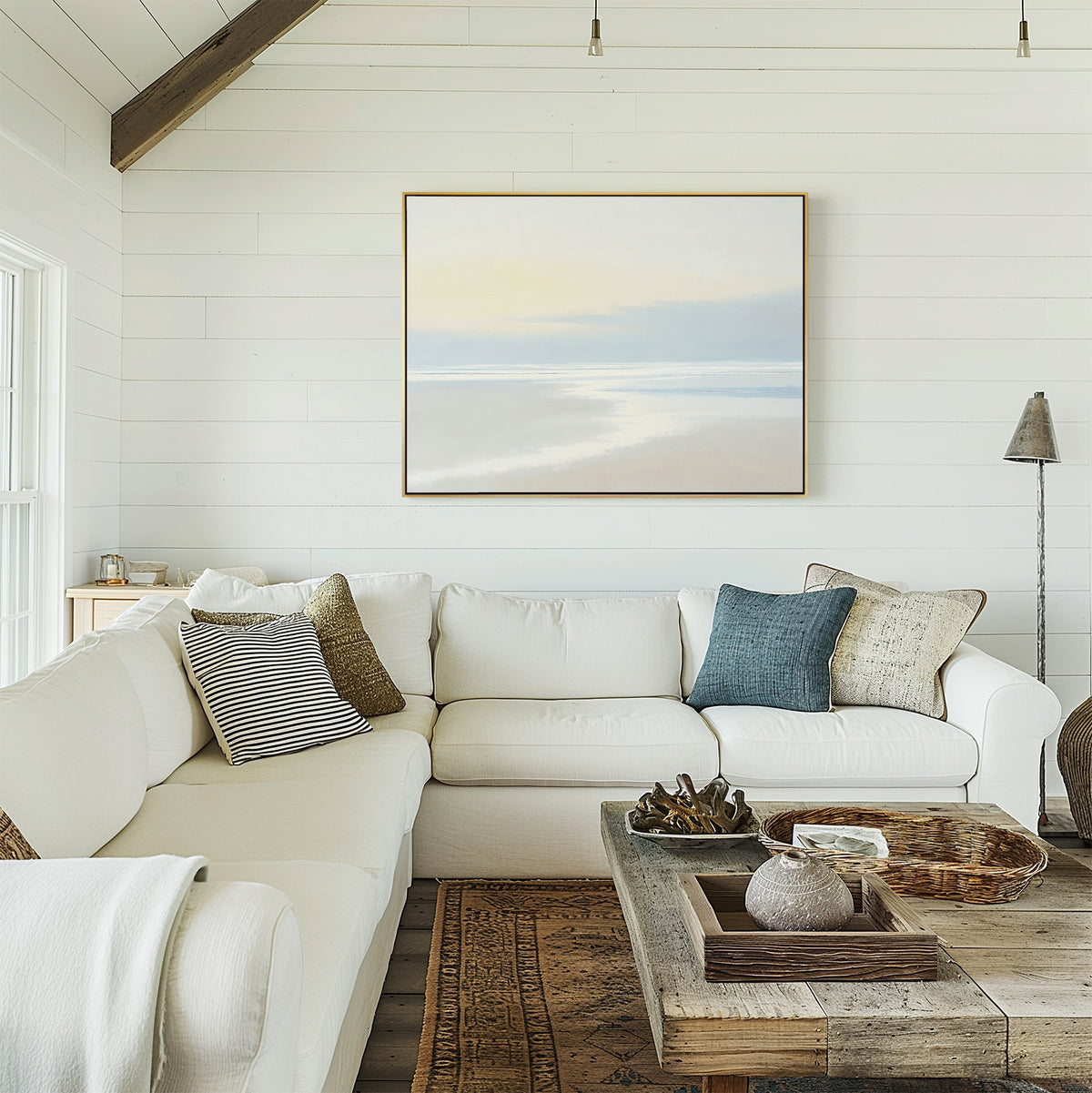 A bright, coastal-style living room with white shiplap walls, a large white sectional sofa, neutral and blue pillows, a rustic wooden coffee table, and the Quiet Shores Canvas Art – Horizontal displayed above the couch.