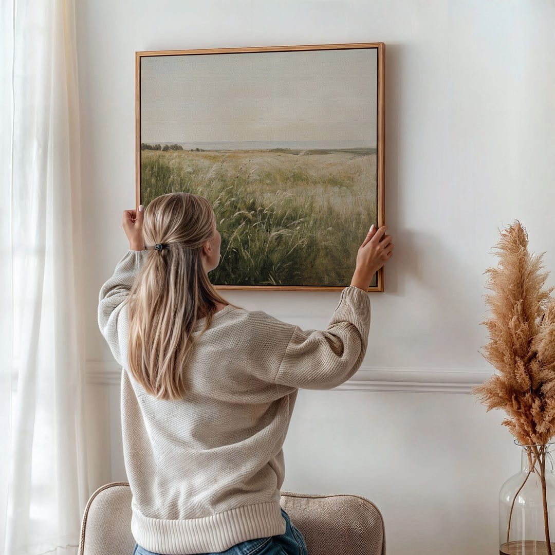 A woman with long blonde ponytail hangs the Reeds by the River Canvas Art on a white wall beside a vase of dried pampas grass, bringing charm to her softly lit living room decor.