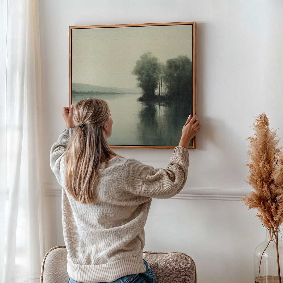 A woman with blonde hair in a ponytail hangs the Reflective River Canvas Art, featuring trees and a lake, on a white wall in a softly lit room with pampas grass in a vase nearby.