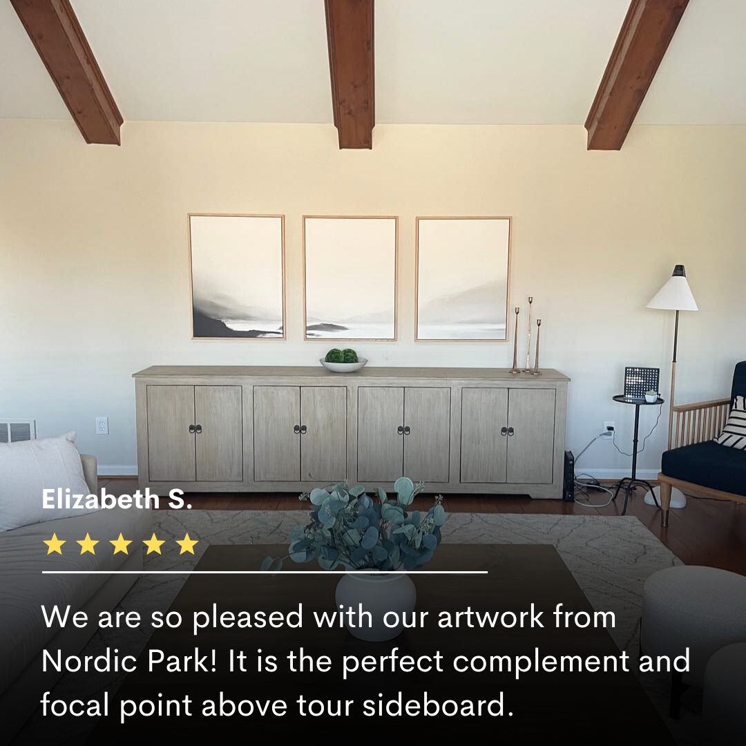 A light wood sideboard sits against a cream wall, topped with minimal decor. Above, the Serenity Peaks Set of 3 abstract landscape prints are evenly spaced. Exposed wooden beams and neutral modern furnishings create a serene balance.