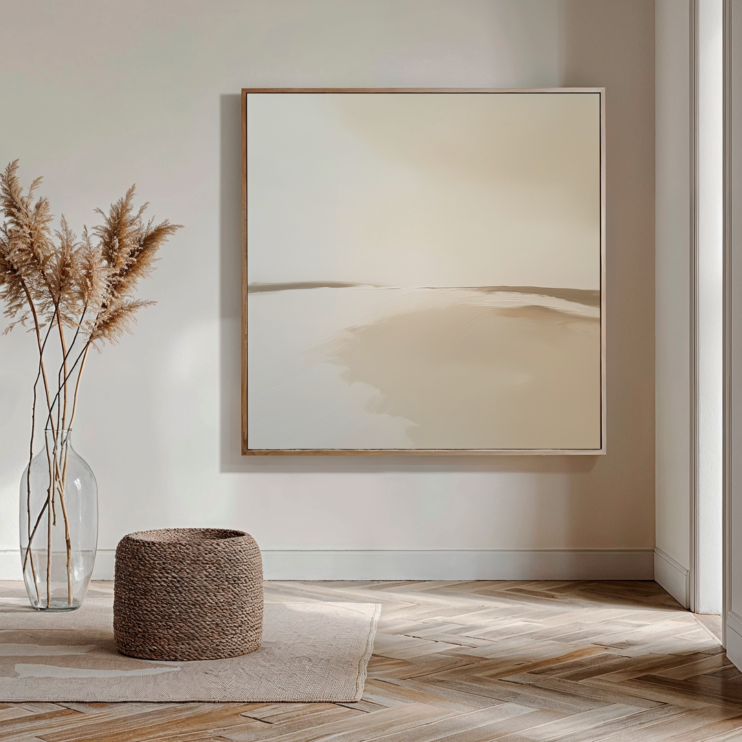A minimalist living room with light walls, My Stores Serene Expanse Abstract Canvas Art in beige tones, a glass vase of dried pampas grass, a woven pouf, and a light rug on herringbone wood flooring. Sunlight enters from the right.