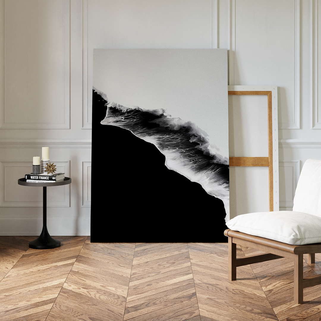 A modern room with chevron wood flooring features the Black Ocean Abstract Canvas Art propped against a wall in a wooden frame, alongside a white cushioned chair and a small black table with candles and books.
