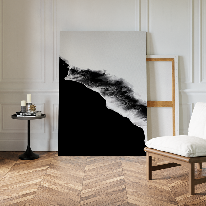 A modern room with chevron wood flooring features the Black Ocean Abstract Canvas Art propped against a wall in a wooden frame, alongside a white cushioned chair and a small black table with candles and books.