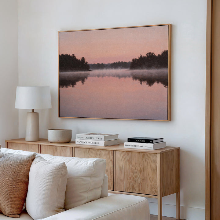 A modern living room features a wooden sideboard, stacked books, a white lamp, and Rose Lake Canvas Art—a framed misty lake landscape at sunset. A white sofa is partially visible, inspiring serene bedroom decor ideas.