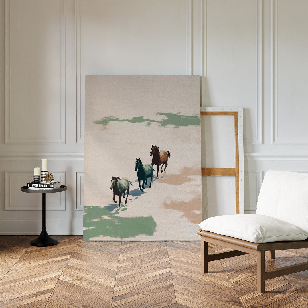 A minimalist room showcases the Running Free Canvas Art of three horses leaning against a white paneled wall, beside a white cushioned chair and a small black round table with candles and books on the herringbone wood floor.