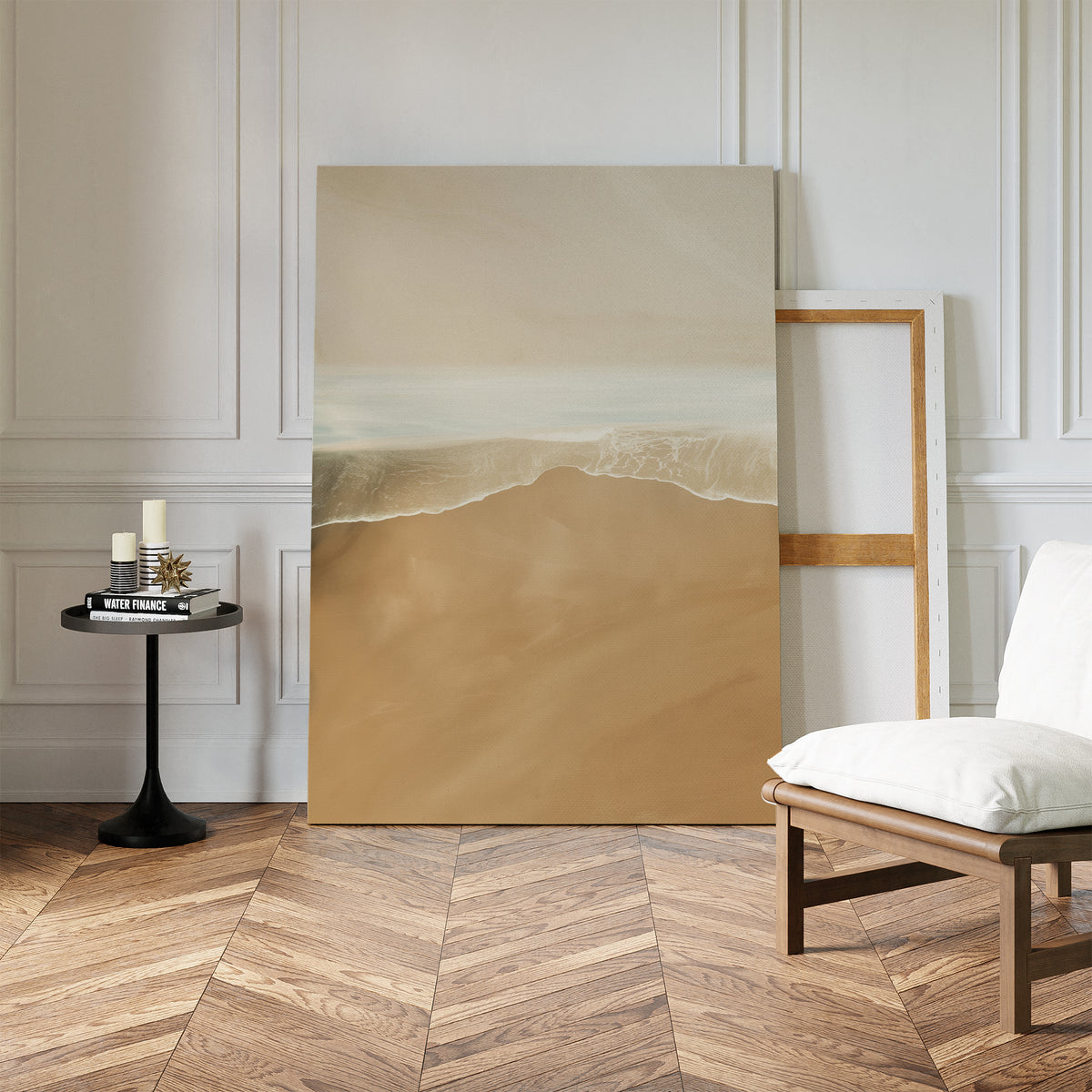 The Sand and Sea Abstract Canvas Art, depicting a serene beach with gentle waves, leans against a white-paneled wall beside an empty wooden frame, a modern cushioned chair, and a round table—ideal for neutral home decor.