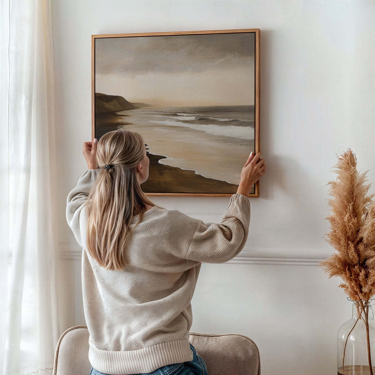 A woman with long blonde hair hangs the Sandy Shoreline Canvas Art, featuring a coastal seascape with cliffs and waves, on a white wall beside a vase of dried pampas grass.
