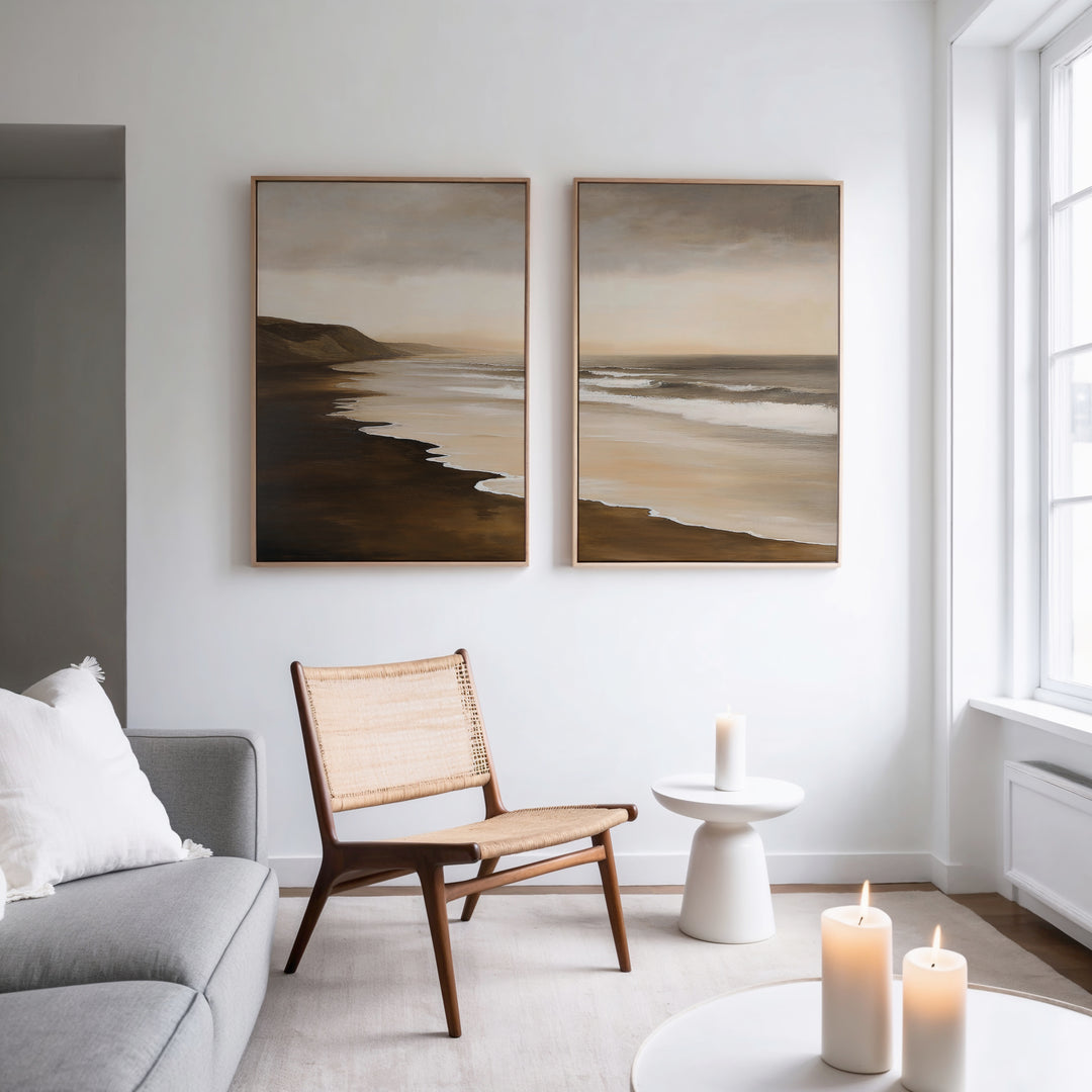 A modern living room with a wooden chair, small white table, lit candles, gray sofa with a white pillow, and the Sandy Shoreline Set of 2 beach canvases hanging above. Natural light pours in from tall windows.