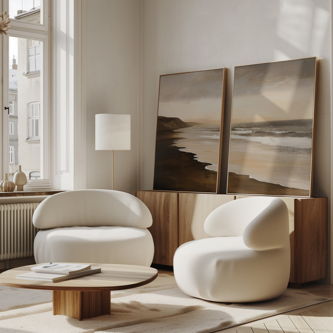 A modern living room features two rounded white armchairs, a low wooden coffee table, and the Sandy Shoreline Set of 2 coastal wall art above a wooden sideboard. Sunlight streams in through tall windows, creating a warm and airy atmosphere.