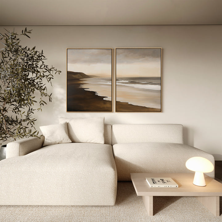 A modern living room features a cream sectional, small wooden table with round lamp and book, potted plant, and the Sandy Shoreline Set of 2 coastal wall art above the sofa for a serene decor touch.