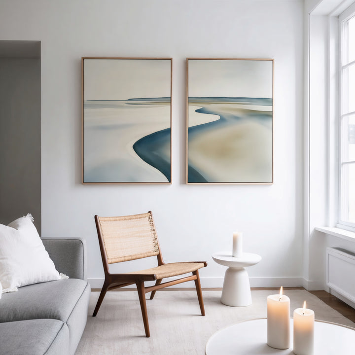 A minimalist living room with a wicker chair, small white table, lit candles, gray sofa, and the Serene Coastline Set of 2 wall art hanging on a white wall near a large window.