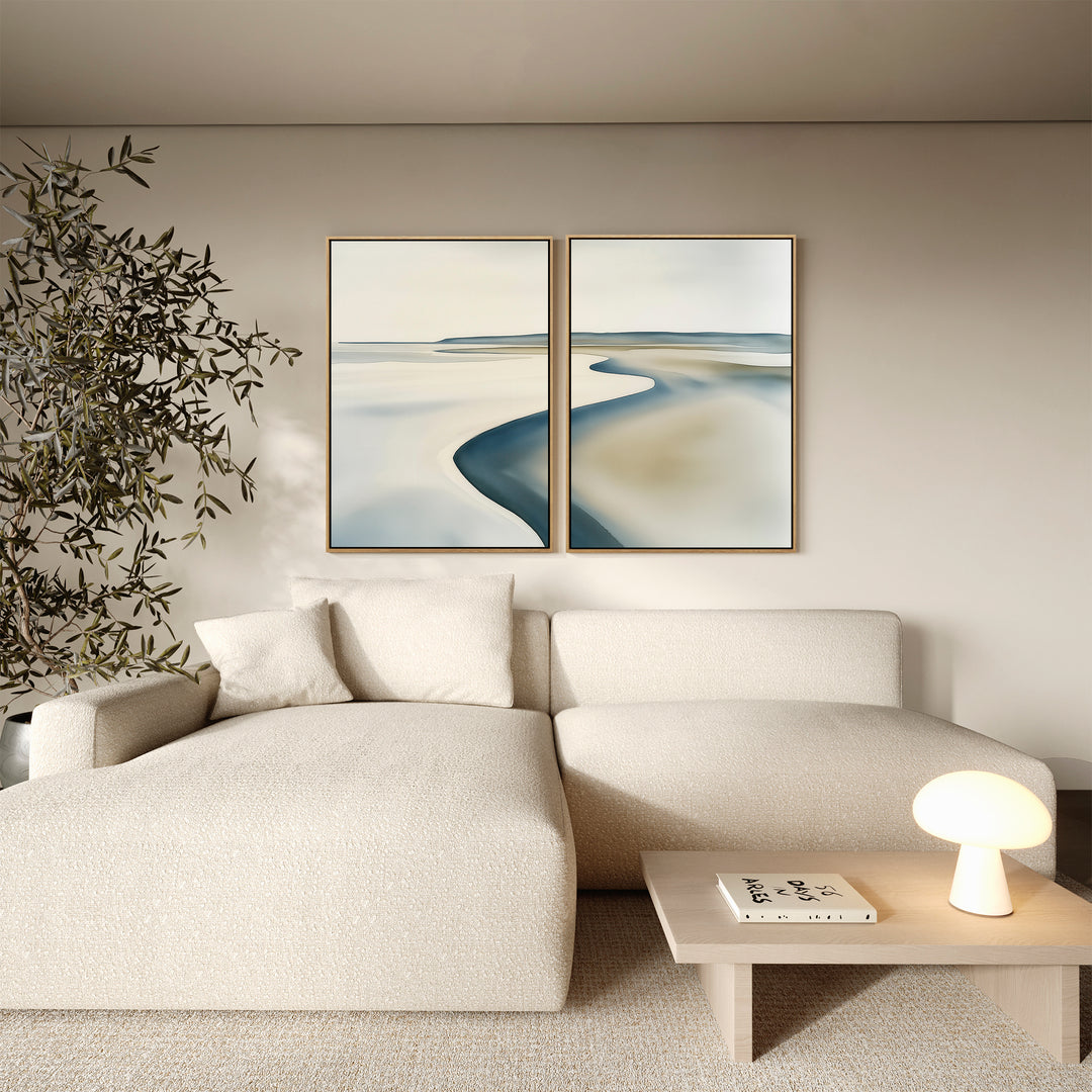 A modern living room with a cream sectional sofa, a wooden coffee table holding a small round lamp and book, an indoor plant, and the Serene Coastline Set of 2 minimalist wall art on a beige wall.