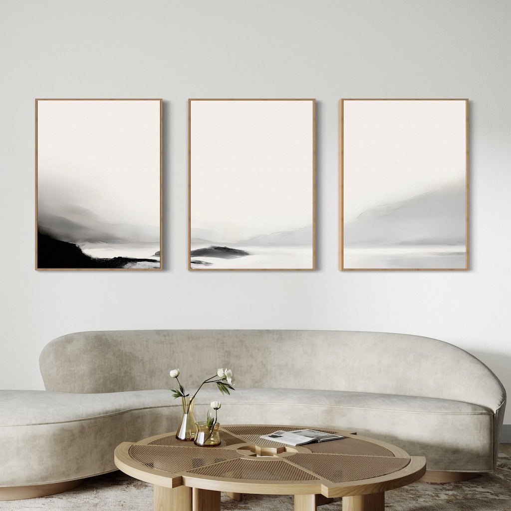 A modern living room featuring a curved beige sofa, a round wooden coffee table with flowers and books, and the Serenity Peaks Set of 3 minimalist landscape paintings in neutral tones creates a relaxed, contemporary space perfect for unwinding.