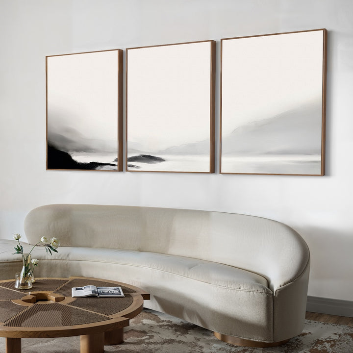A modern living room features a curved beige sofa, a round wooden coffee table, and the Serenity Peaks Set of 3 black-and-white abstract landscape paintings above. A vase with flowers adds freshness to the serene, contemporary space.