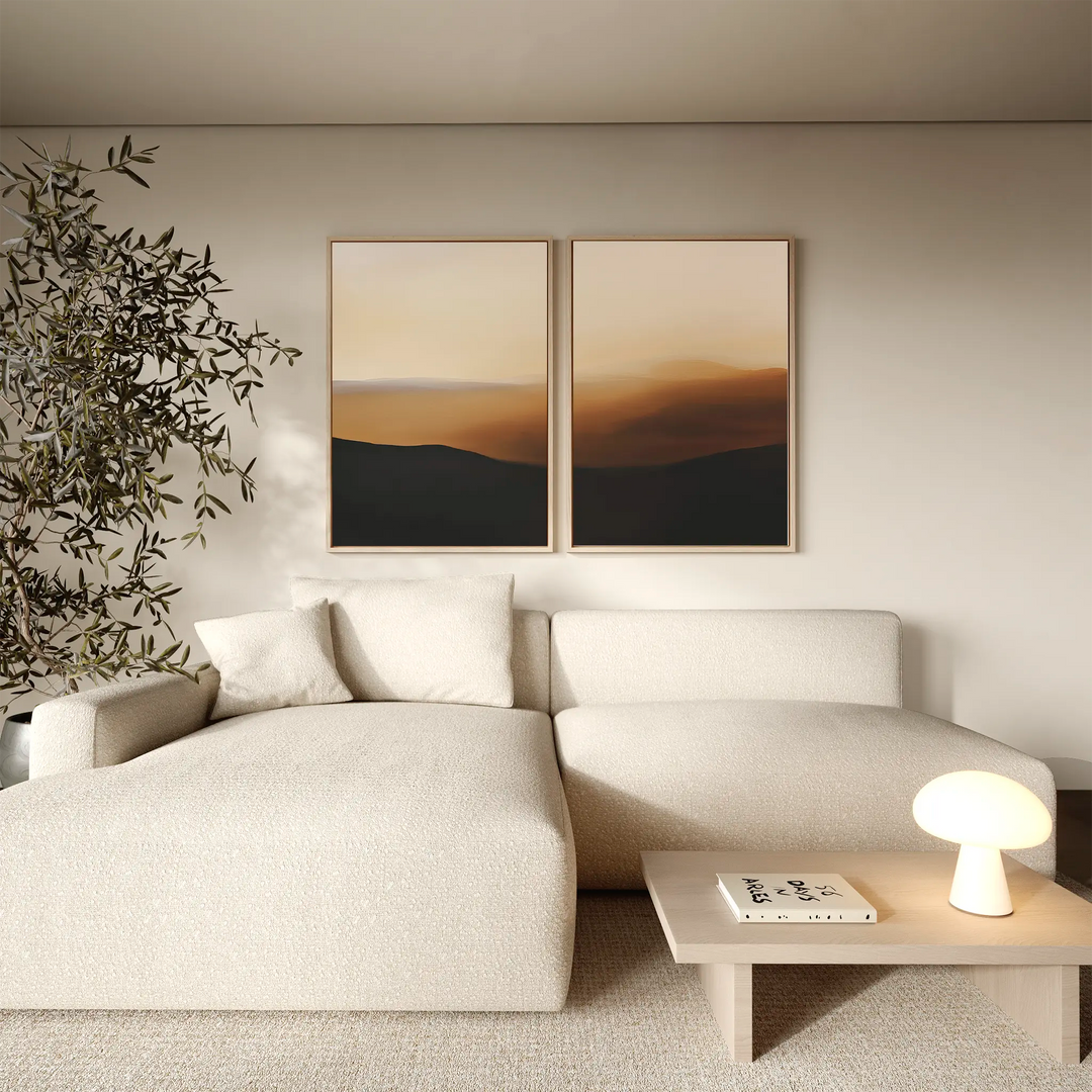 A minimalist living room with a cream sofa, small wooden coffee table holding a lamp and book, two Sienna Echo Set of 2 wall art pieces above, and a leafy plant in the corner.