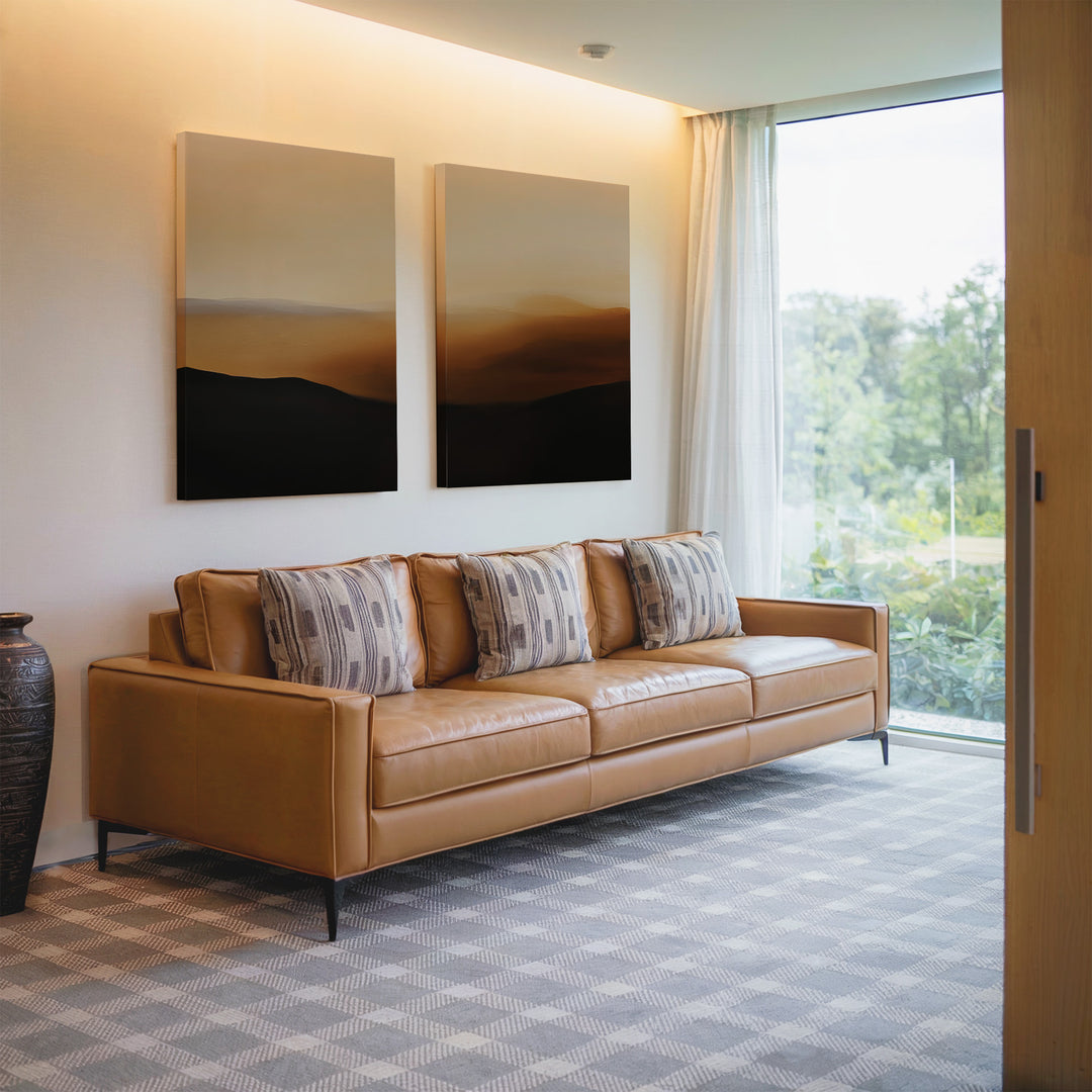 A modern living room featuring the Sienna Echo Set of 2 tan leather sofas, four patterned cushions, minimalist landscape wall art above, a large ceramic vase, and a window with sheer curtains overlooking greenery—stylish decor.